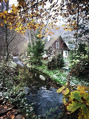 Une maison au bord d’une rivière, avec une terrasse en bois et une montagne en arrière-plan.