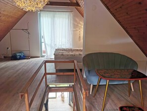 A loft bedroom with a sloped ceiling, a bed with a floral bedspread, a green armchair, a small table with a floral pattern, and a wooden bench. - Fairy Tale Cottage on Water Stream, 30 Minutes From South Prague (Petrov)