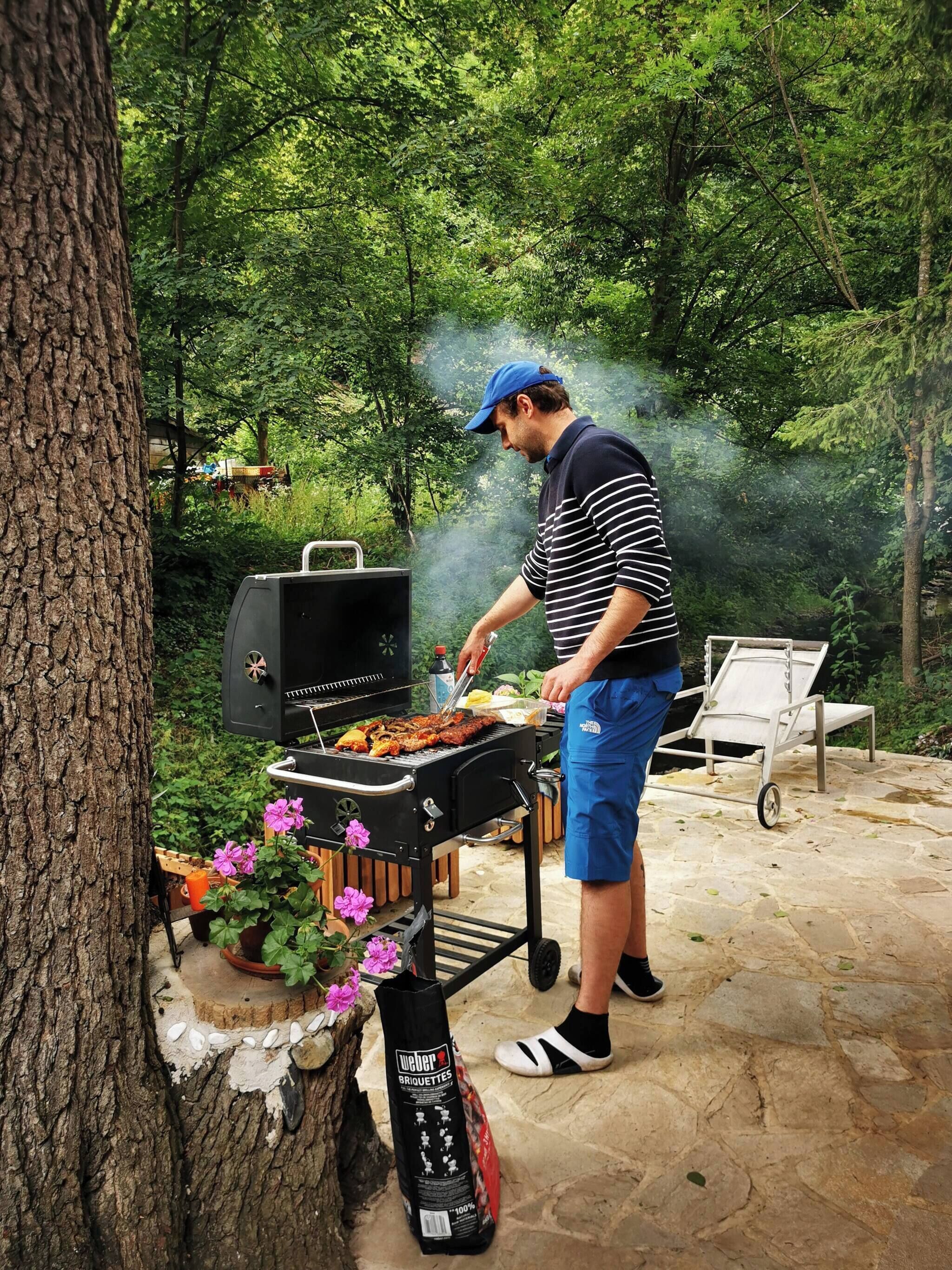 Un uomo cucina all'aperto con una griglia portatile, circondato da vegetazione e un patio in pietra.