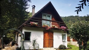 Una casa de dos pisos con techo inclinado, balcón de madera con jardineras y una escalera de piedra que lleva a la entrada.