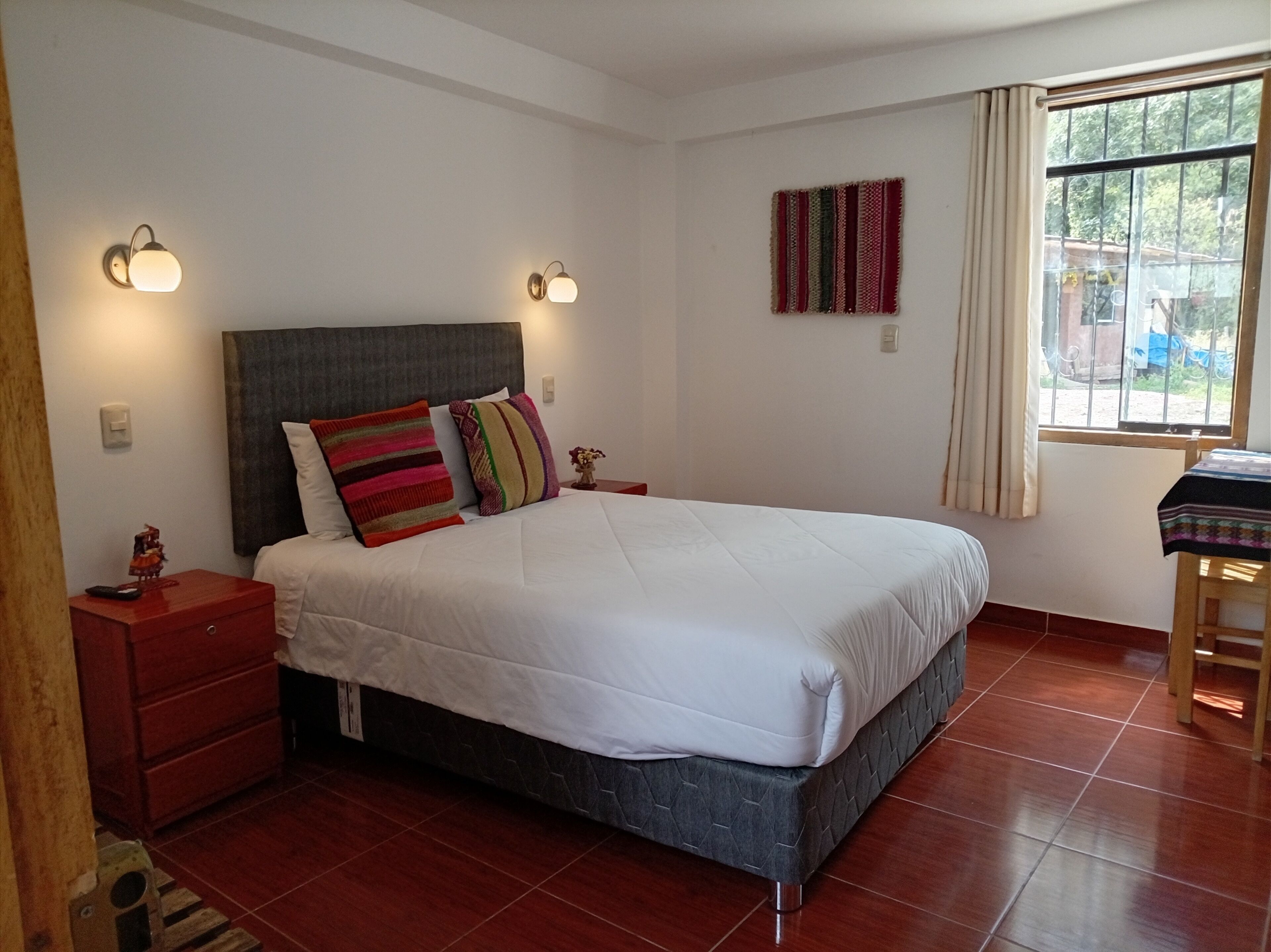 A bedroom with a large bed, two bedside tables, a nightstand, a window with curtains, and a wall-mounted artwork.
