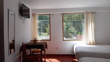Habitación con cama, escritorio y silla, televisor montado en la pared y dos ventanas con cortinas.