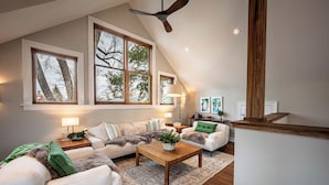 A spacious living room with a high ceiling, large windows, and a wooden ceiling fan.