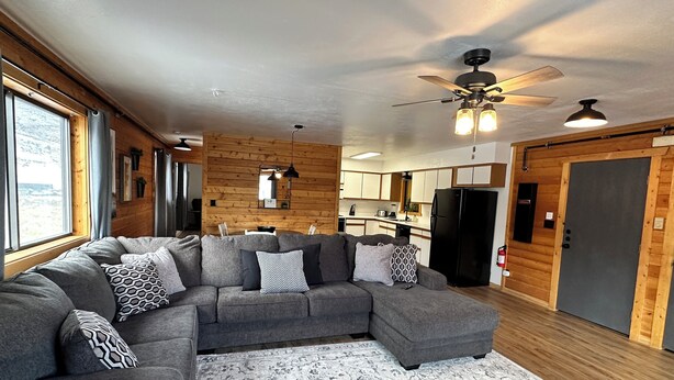 A modern living room with a gray sectional sofa, a patterned rug, wooden walls, a kitchen area, and a ceiling fan. - Cozy 1-bedroom apartment unit C, sleeps 6  across from famous Cody Nite Rodeo! (Cody)