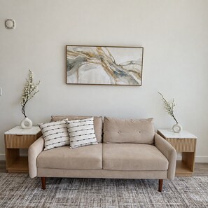 Ein neutral eingerichtetes Wohnzimmer mit einem beige farbenen Sofa, zwei Beistelltischen mit Vasen und einem großen abstrakten Gemälde an der Wand.