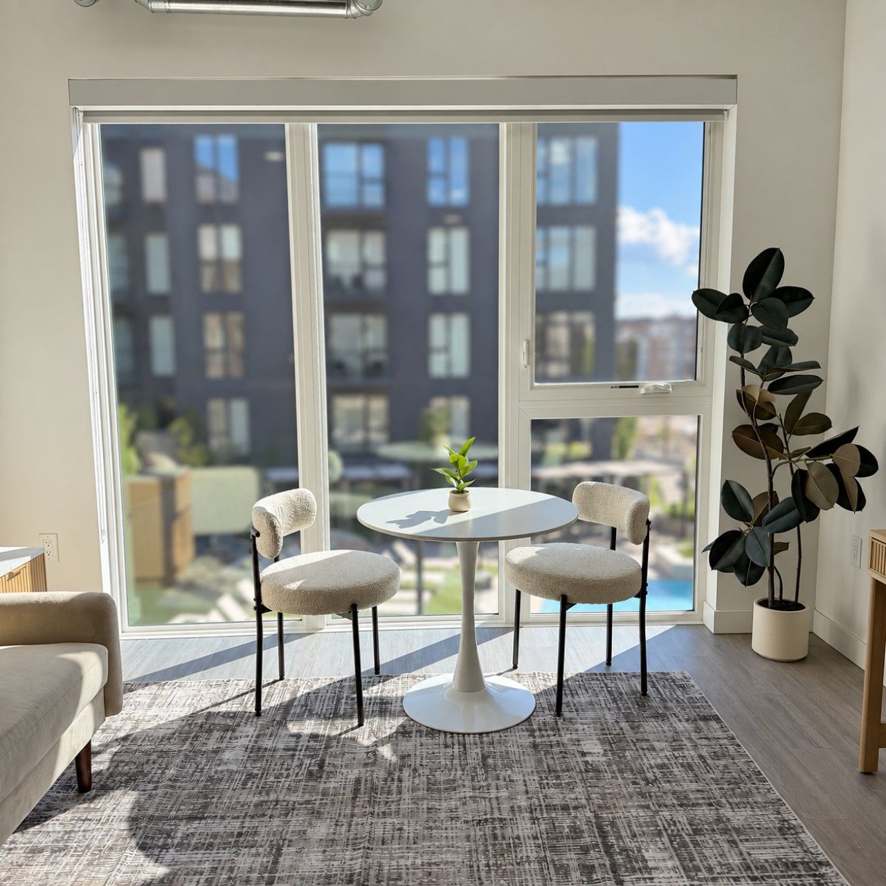 A room with a round table, four chairs, a potted plant, and a view of buildings through large windows.
