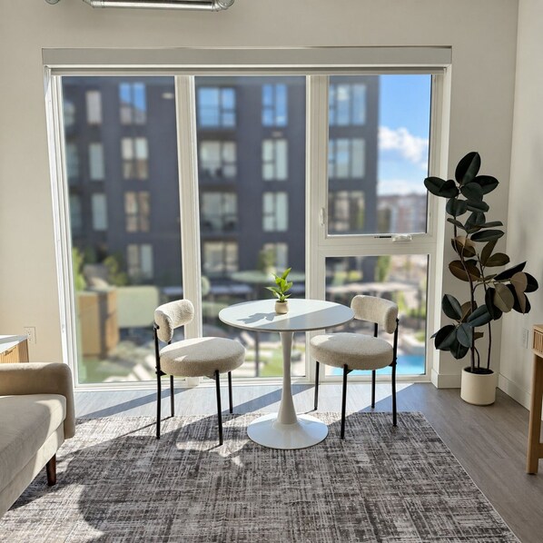 A room with a round table, four chairs, a potted plant, and a view of buildings through large windows.