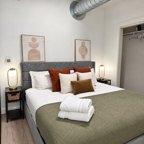 Ein ordentlich bezogenes Bett mit grauem Kopfteil, weißen und braunen Kissen und einer grünen Tagesdecke. Zwei Nachttische mit Lampen stehen daneben, und an der Wand über dem Bett ist Kunst ausgestellt.