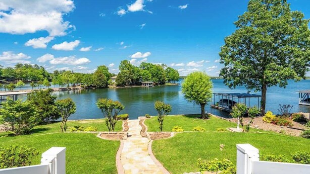 A lakeside property with a stone pathway leading to a dock, surrounded by lush greenery and a clear blue sky.