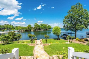Una propiedad junto al lago con un camino de piedra que lleva a un muelle, rodeada de exuberante vegetación y un cielo azul despejado.