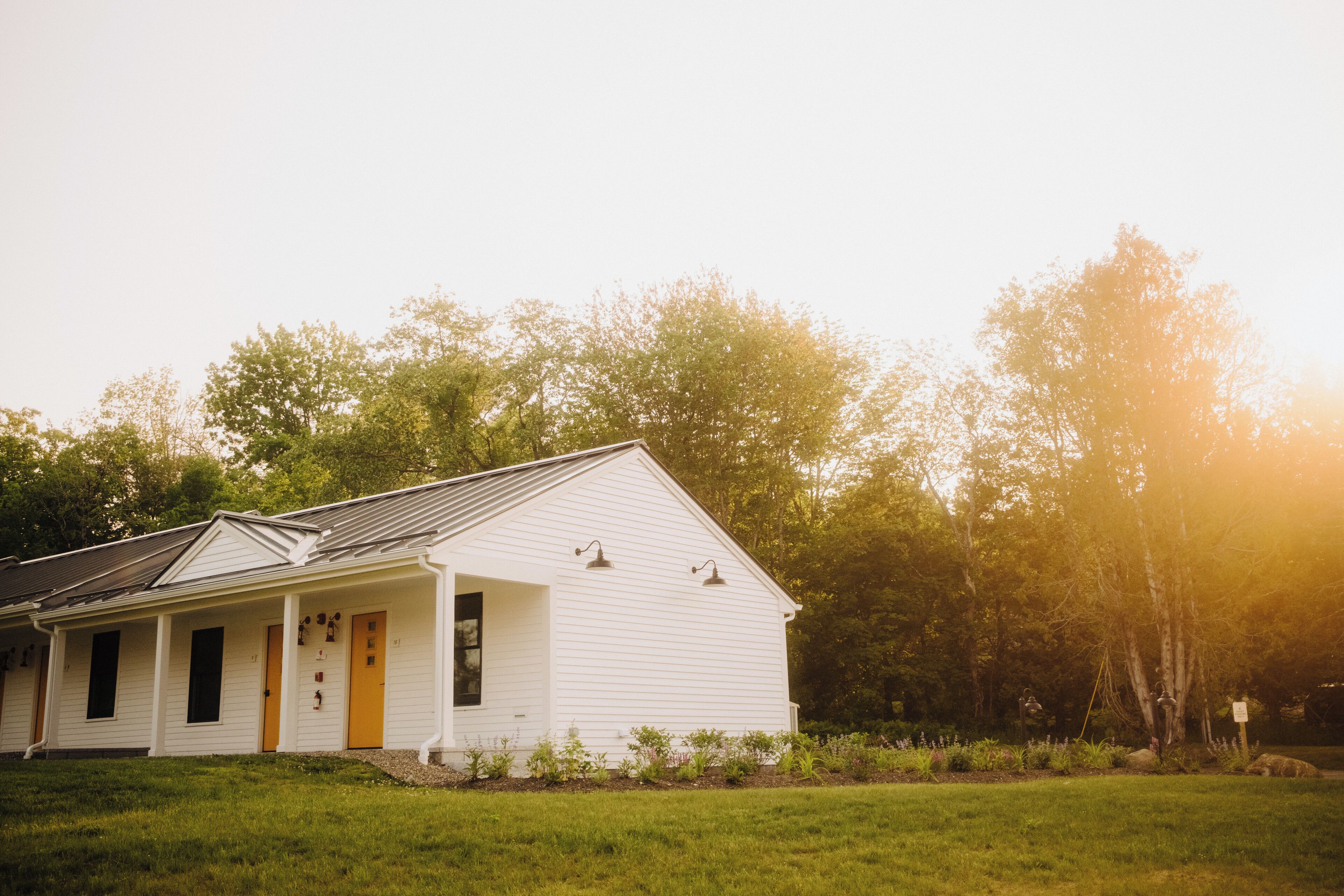 A white building with a porch and a yellow door, surrounded by greenery.