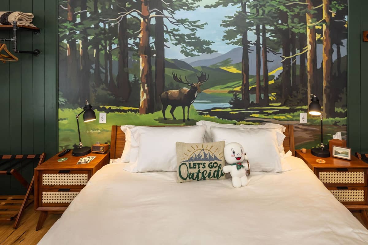 A bedroom with a bed, two nightstands, a lamp, a plush toy, and a wall mural of a forest scene.