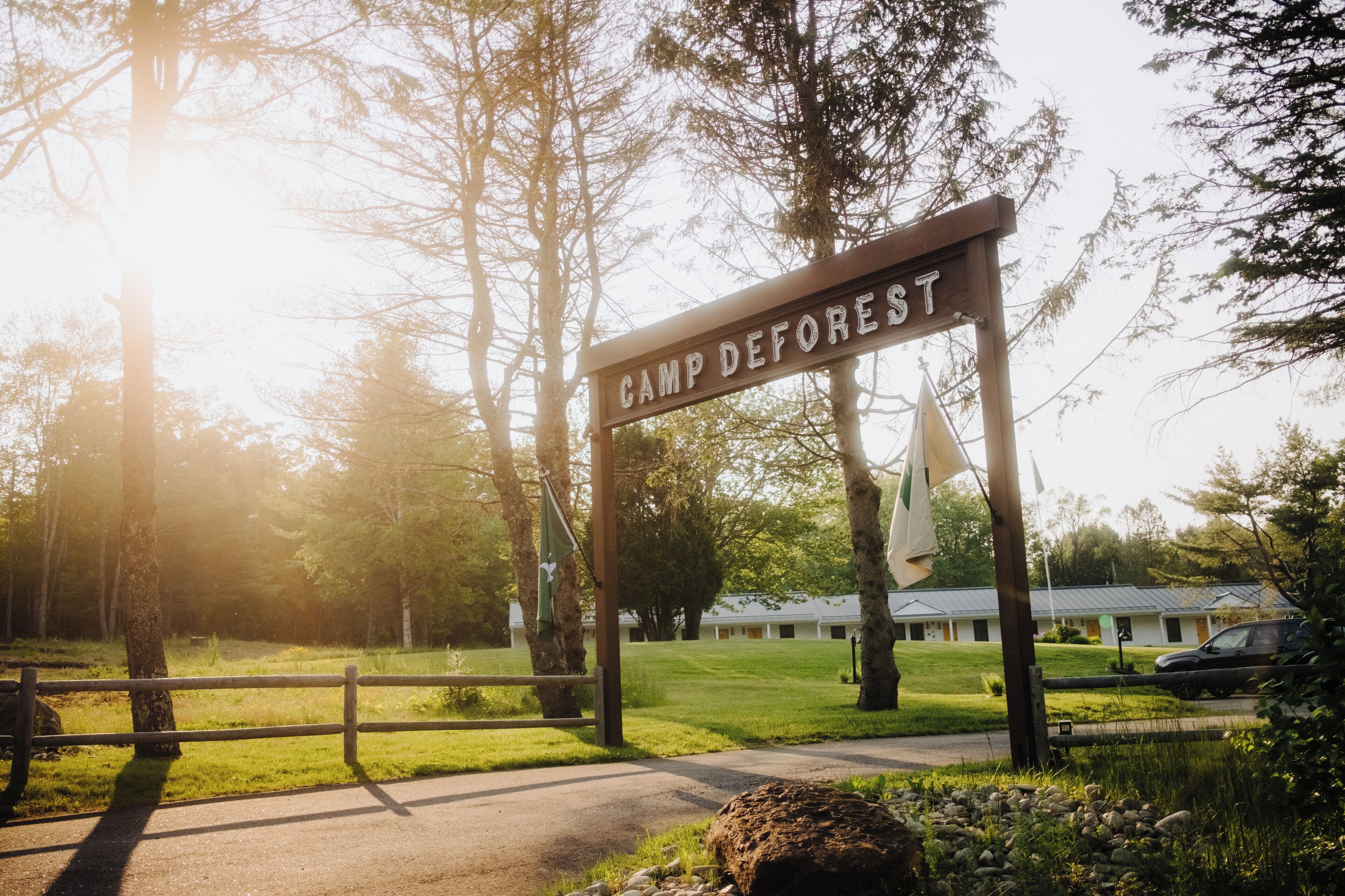 A wooden sign with the text "CAMP DEFOREST" in a natural setting with trees and a building in the background.