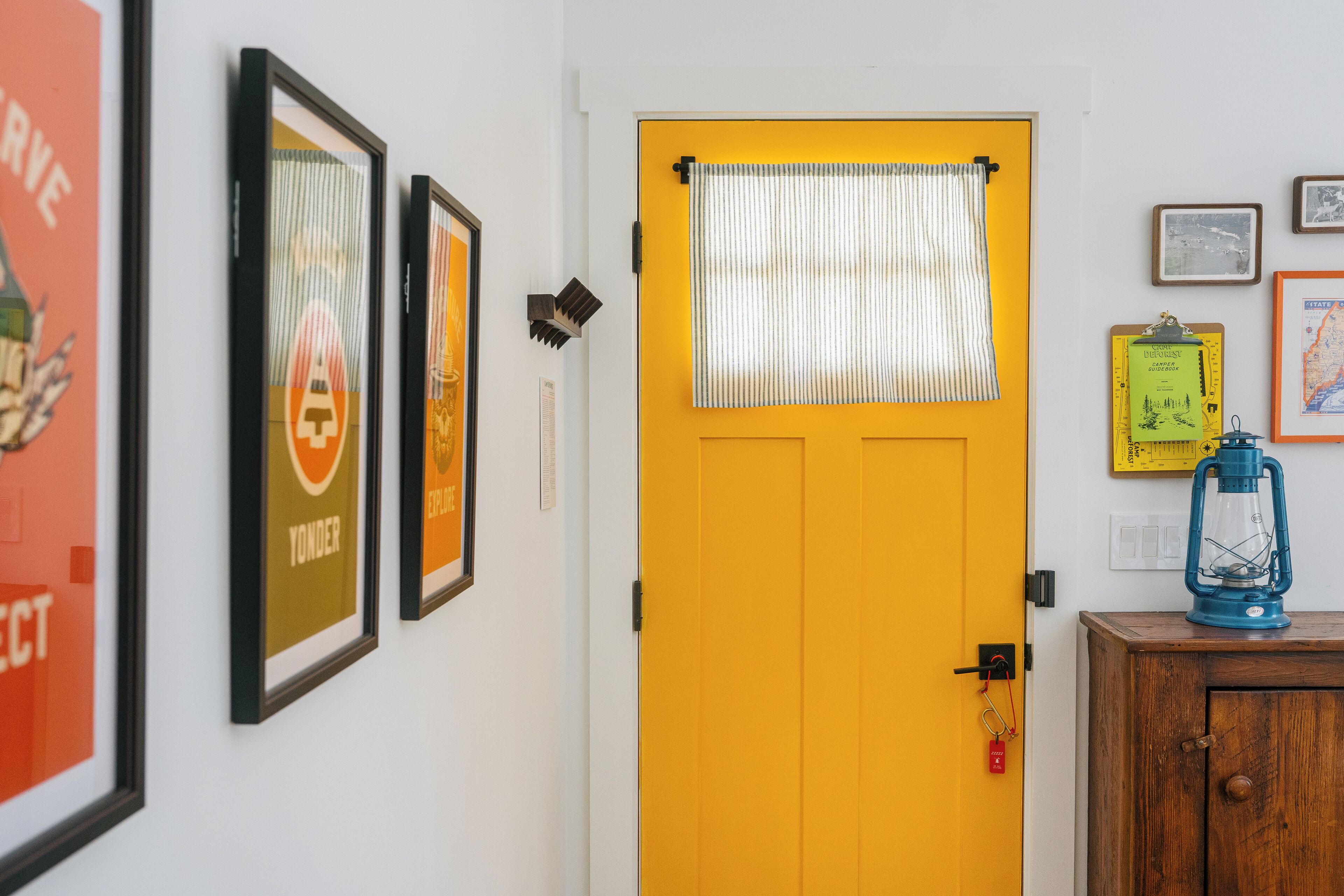 Una puerta amarilla con una cortina a rayas, carteles enmarcados en la pared, un mueble de madera con un farolillo y un llavero pequeño.