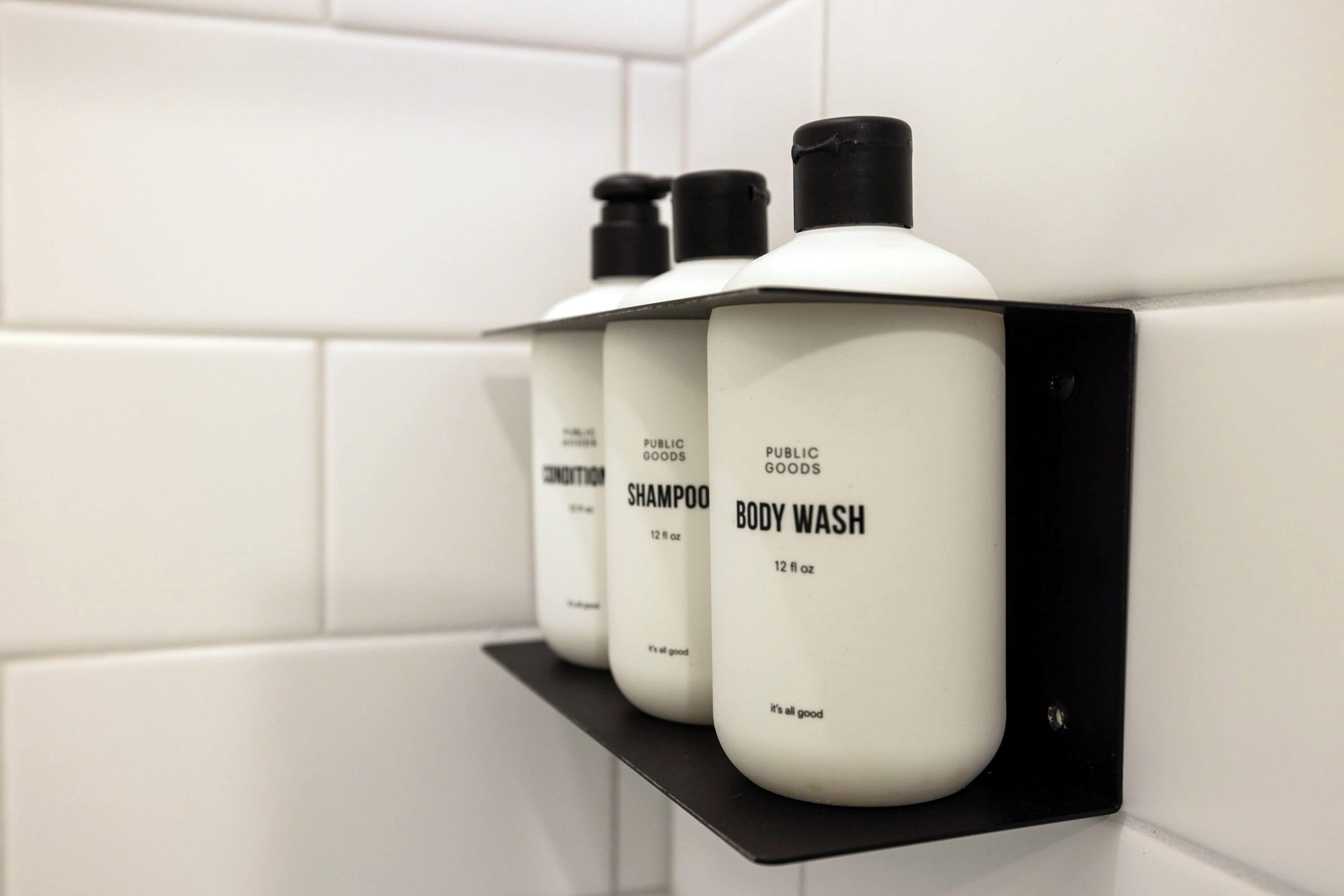 Three bottles of body wash and shampoo on a black shelf in a bathroom with white tiles.