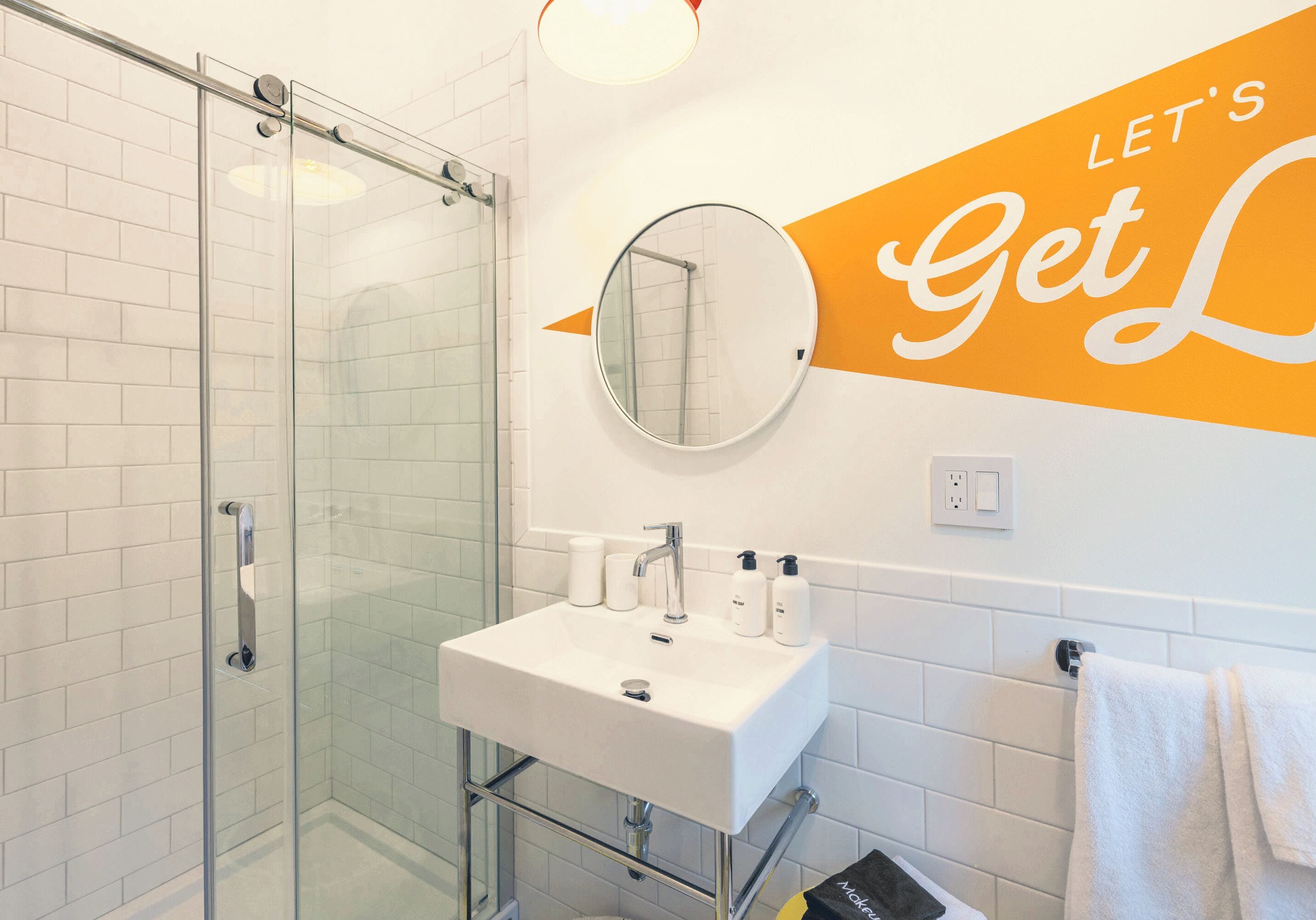 A modern bathroom with a glass shower enclosure, a white sink with toiletries, and a motivational sign on the wall.