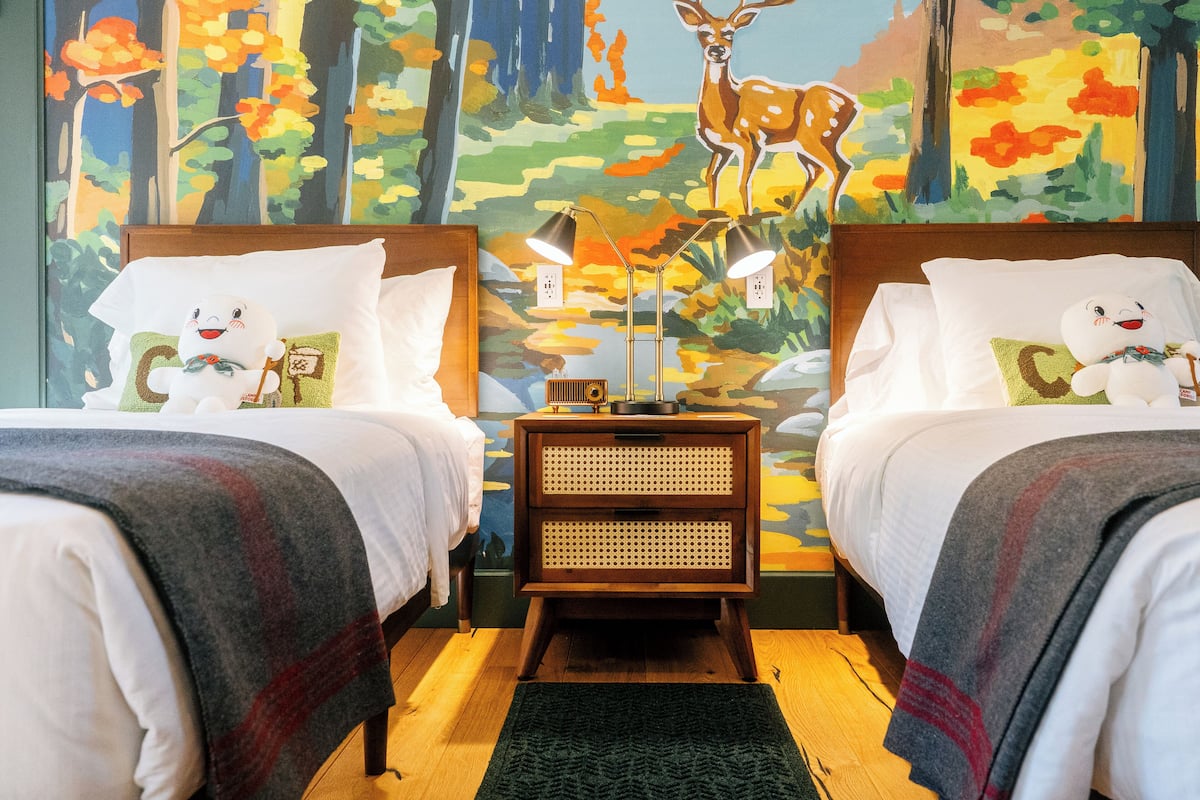 A hotel room with two beds, a nightstand with lamps, a decorative wall mural, and a small table with a lamp.