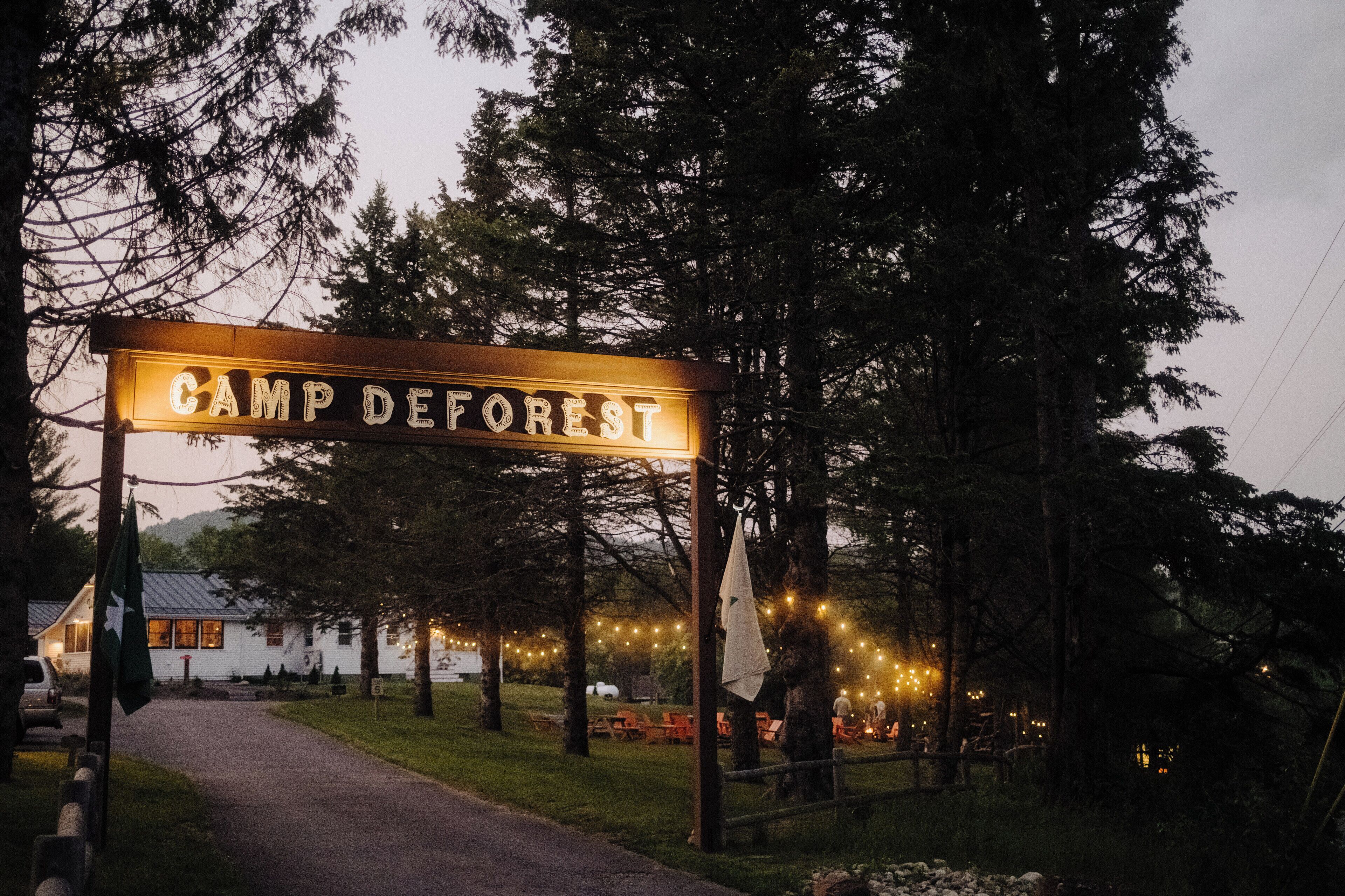Une pancarte indiquant « Camp DeForest », surmontée d’une arche rustique en bois, entourée d’arbres et d’un sentier menant à un bâtiment illuminé de guirlandes lumineuses.