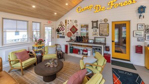 A cozy living room with a wooden ceiling, a neon sign reading "Camp De Forest," and a variety of furniture including a coffee table, chairs, and a bookshelf. - Camp DeForest (Lincolnville)