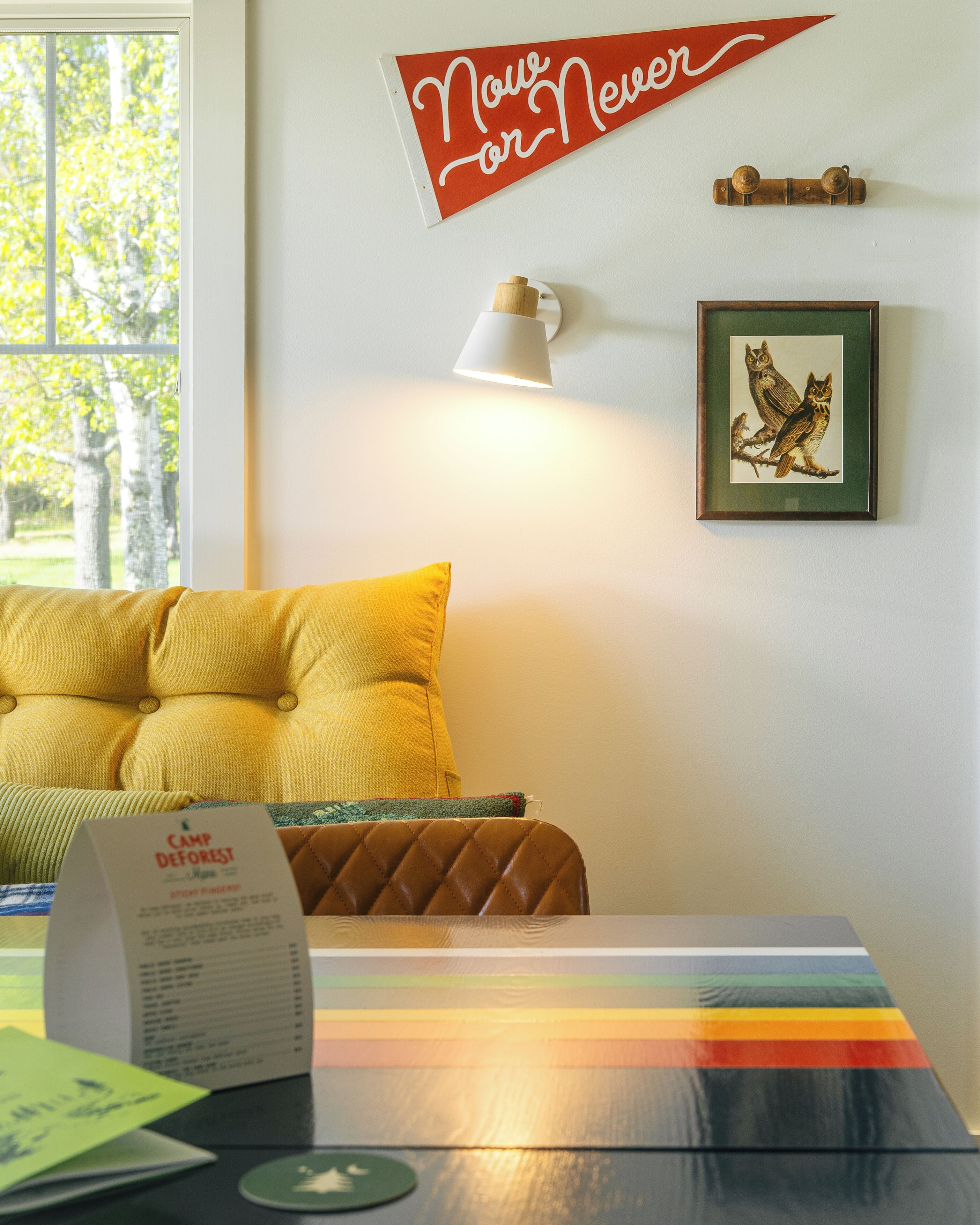 A room with a yellow pillow, a framed picture of owls, a 'Now or Never' sign, and a lamp.
