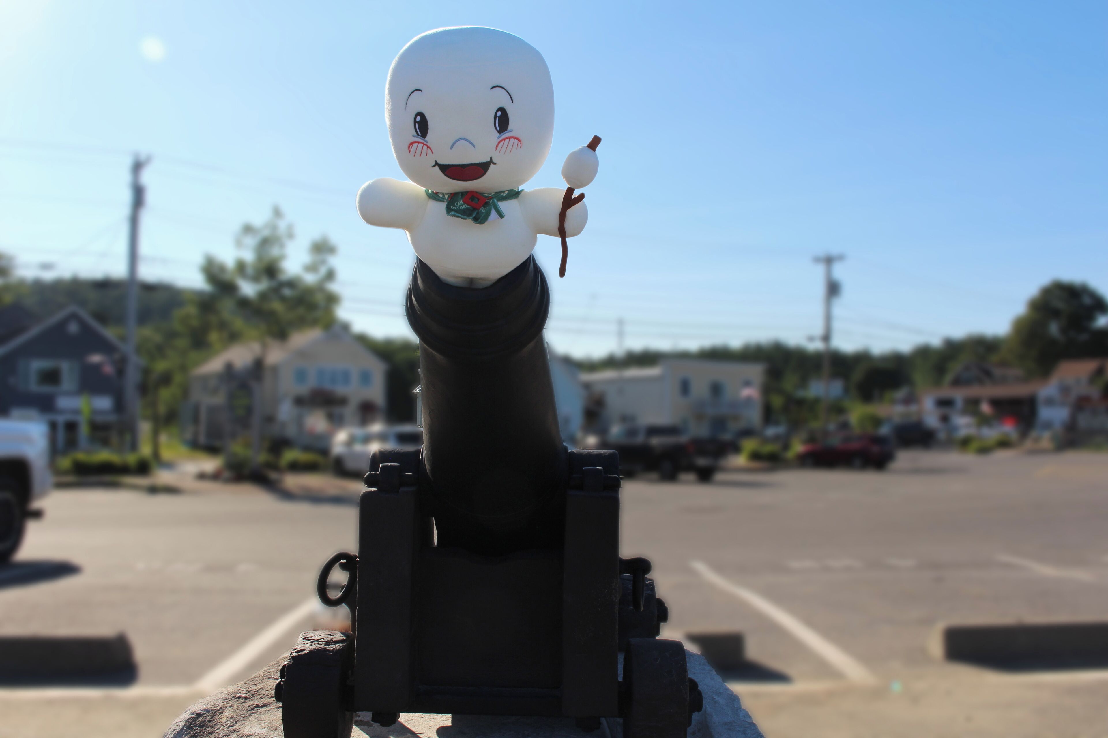 A figurine of a character with a green scarf and a stick, placed on a black stand in a parking lot with houses and cars in the background.