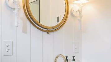 Un baño con tocador blanco, espejo con marco dorado y dos apliques de luz.
