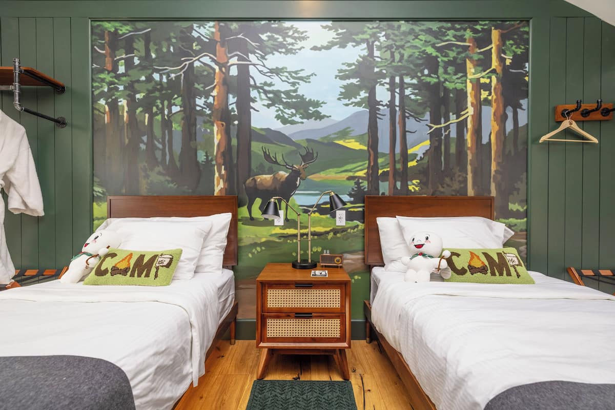 A room with two beds, a nightstand with a lamp, and a large forest mural on the wall.