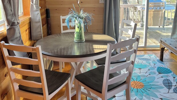 L'intérieur d'un chalet en bois, avec une table à manger ronde, des chaises blanches, un téléviseur fixé au mur et une vue sur l'extérieur.