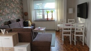 Una sala de estar con un sofá, una mesa de comedor con sillas, un televisor y una ventana con plantas.