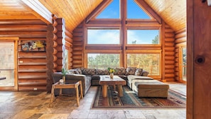 A log cabin interior with a large window, a sofa, a coffee table, and a rug. - Sleeps 27 2 Homes Near the Lake, Hot Tub + Views Castle Glen Estates (Big Bear Lake)