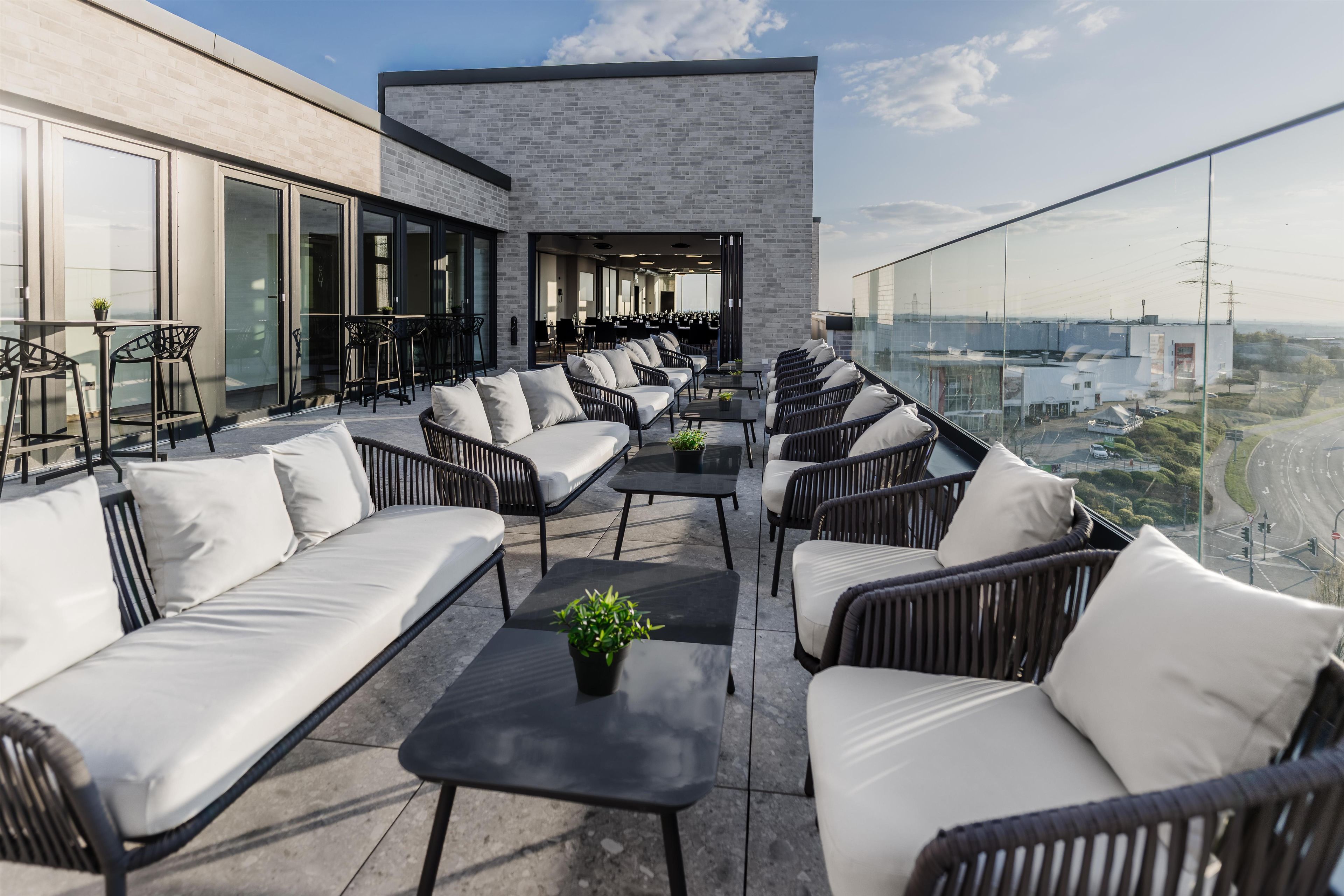 Eine Dachterrasse mit Sitzgelegenheiten im Freien, ein modernes Gebäude mit großen Fenstern und Blick auf die Stadtlandschaft.
