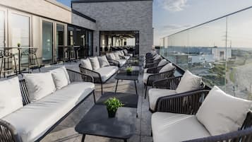 Eine Dachterrasse mit Sitzgelegenheiten im Freien, ein modernes Gebäude mit großen Fenstern und Blick auf die Stadtlandschaft.