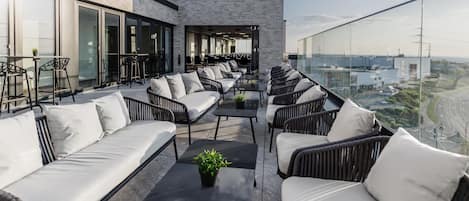 Una terraza en la azotea con mobiliario exterior, un edificio moderno con ventanales grandes y vistas panorámicas de la ciudad.