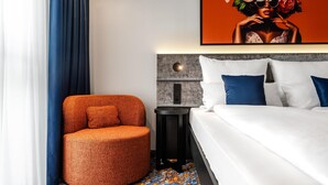 A modern hotel room with a round orange chair, a bed with white and blue bedding, a gray headboard, and a painting above the bed. - Aiden by Best Western Velbert (Velbert)