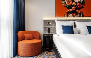 Una camera d'albergo moderna con una poltrona arancione rotonda, un letto con biancheria da letto bianca e blu, un poggiatesta grigio e un quadro sopra il letto.