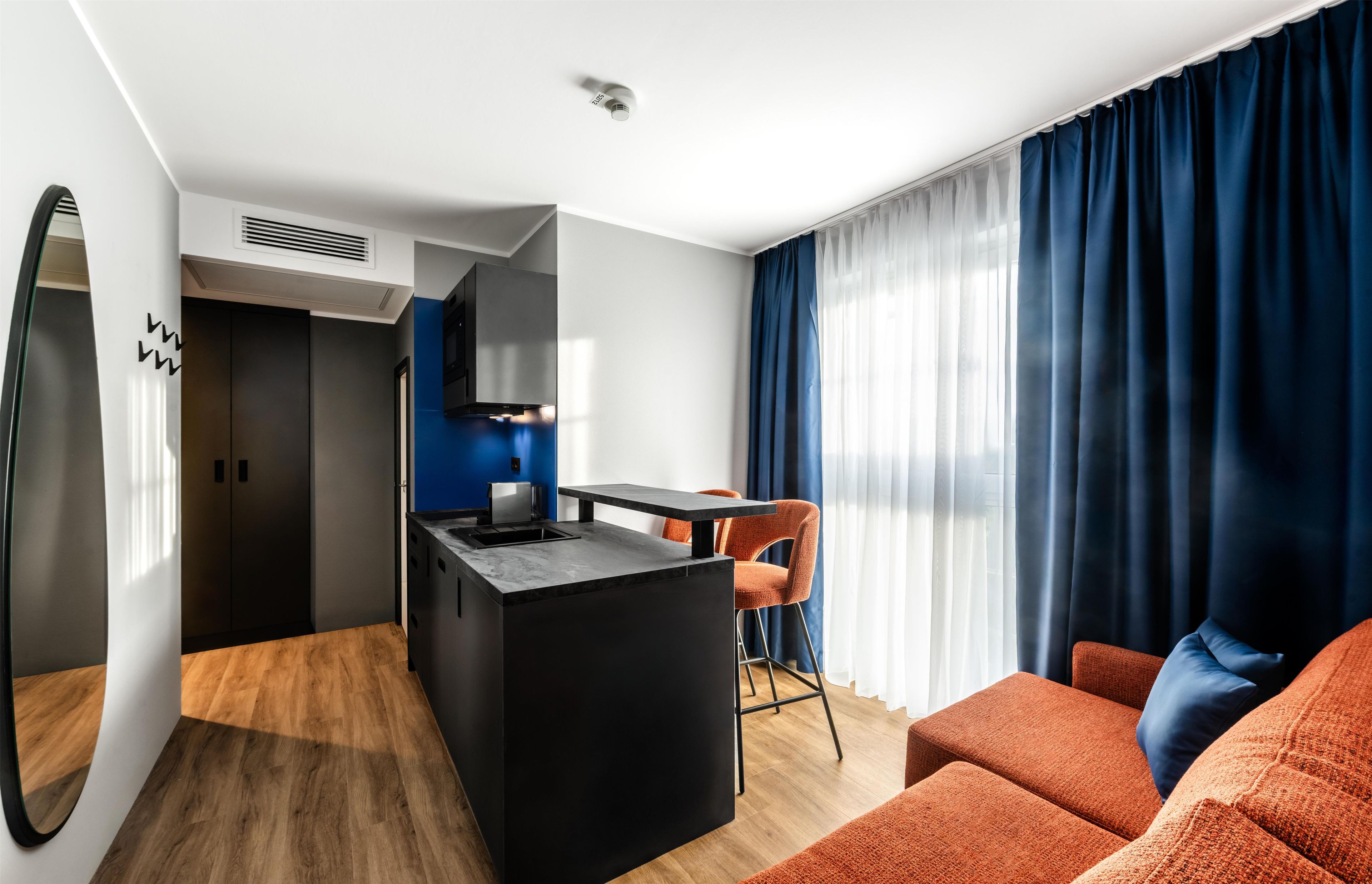 A modern hotel room with a kitchenette, a dining area, and a seating area with a sofa and chair.