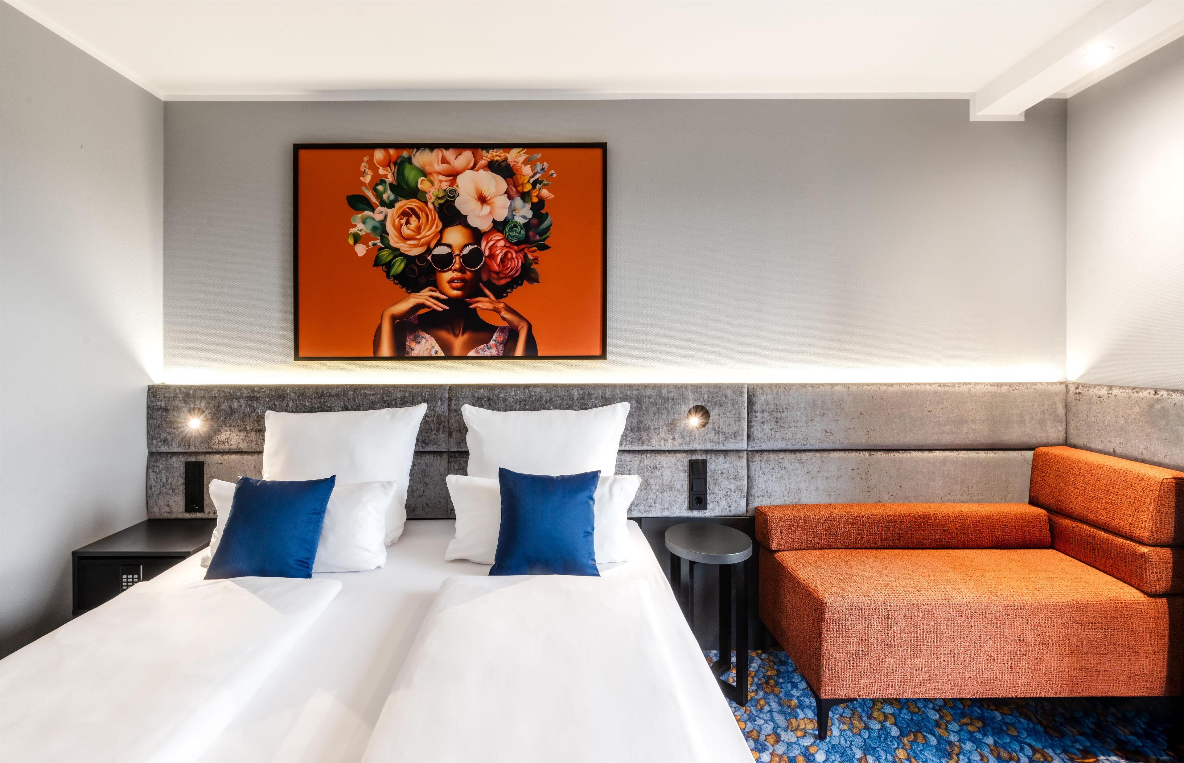 A hotel room with a bed, a blue and white pillow set, a small round table, an orange armchair, and a large floral painting on the wall.