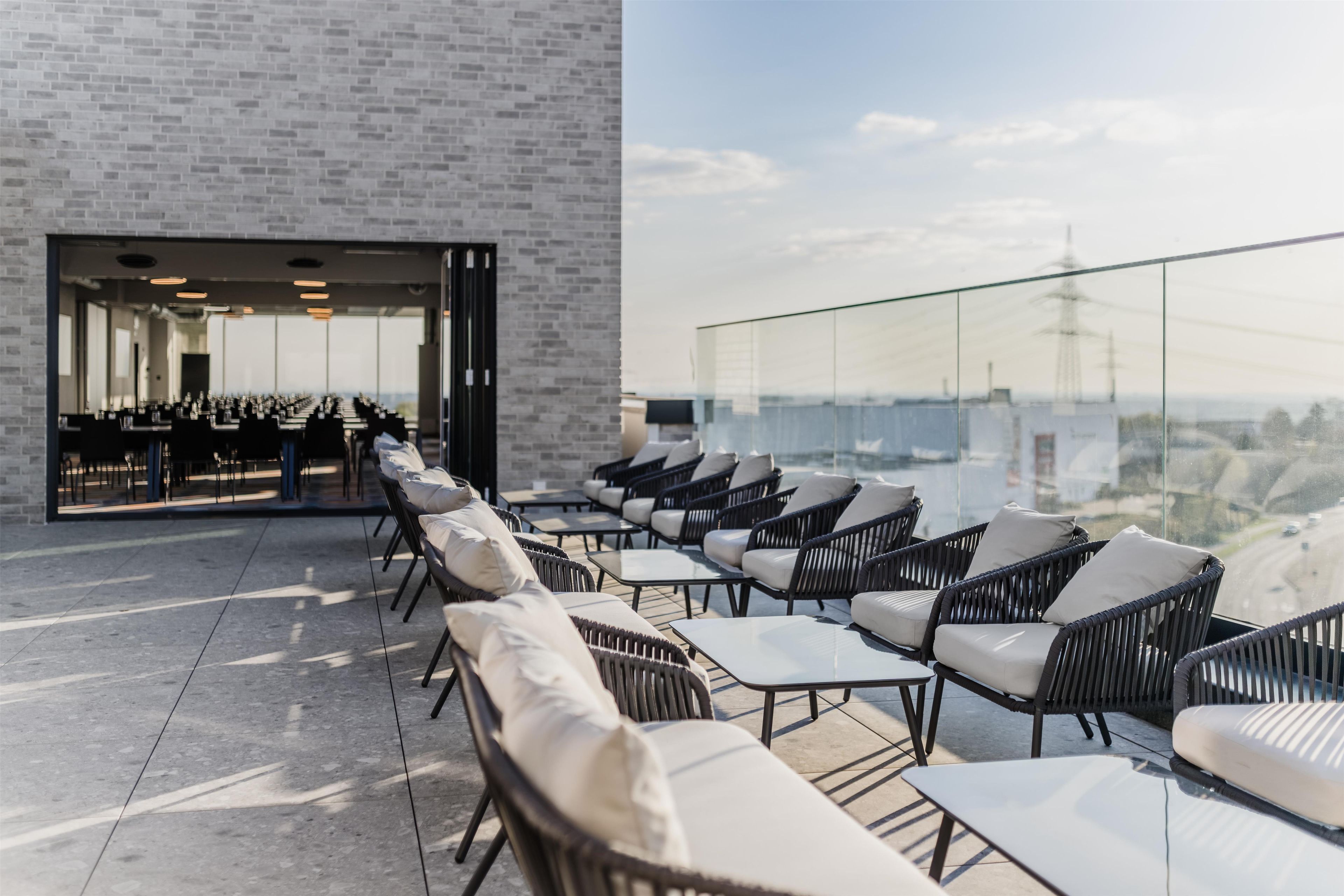 Terraza en la azotea con mobiliario de mimbre y una pared de vidrio que ofrece vistas de la ciudad.