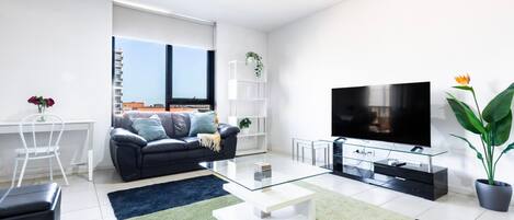 A modern living room with a black leather sofa, a glass coffee table, a flat-screen TV, and a large window offering a city view.