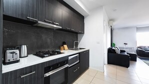A modern kitchen with dark cabinetry, a gas stove, a built-in oven, and a dishwasher. There is a dining area with a black sofa and a white table. - Rowan Place Convenient Home Away With Free Parking (North Melbourne)