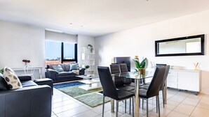 A modern living room with a glass dining table, black leather chairs, a sofa, and a view of the city through large windows. - Rowan Place Convenient Home Away With Free Parking (North Melbourne)