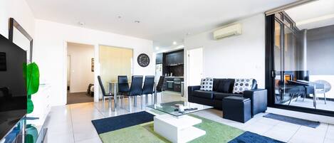 A modern living room with a glass coffee table, black sofa, and a dining area with chairs.