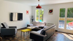 A modern living room with a sofa, armchairs, a TV, a fan, and a window with a view of a pool and greenery. - Family home in Trébeurden close to beaches and hiking trails. (Trébeurden)