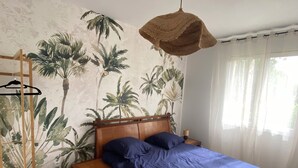 A bedroom with a bed, wooden headboard, a hanging light fixture, a nightstand, a wooden shelf with towels, and a wall with a tropical plant mural. - Family home in Trébeurden close to beaches and hiking trails. (Trébeurden)