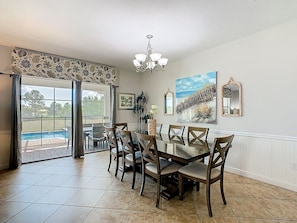 A dining area with a glass table and wooden chairs, a chandelier, and a view of a swimming pool through large windows.
