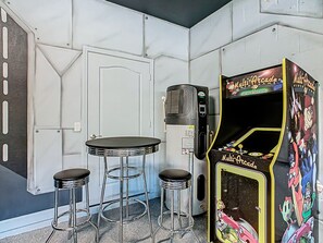 Une pièce comprenant un coin bar avec une borne d’arcade proposant plusieurs jeux vidéo, deux tabourets et une petite table.