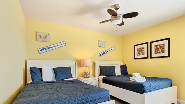 A room with two beds, each with a blue quilted bedspread, white headboards, and a ceiling fan. There are wall-mounted picture frames and a small table with a lamp. - Stunning 8BR Villa with Private Pool & Gameroom (Four Corners)