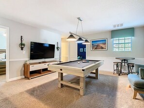 Uma mesa de bilhar numa sala com televisão, área de bar e um quadro na parede.