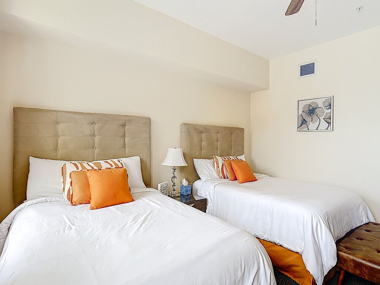 Une chambre d’hôtel avec deux lits, chacun agrémenté de draps blancs et d’oreillers orange. On y trouve une table de chevet avec une lampe, ainsi qu’une petite table sur laquelle reposent une boîte de mouchoirs et un cadre photo.
