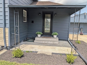 Una casa residencial con fachada gris oscuro, una puerta principal con un panel de vidrio decorativo, dos macetas con plantas en los escalones y un buzón.